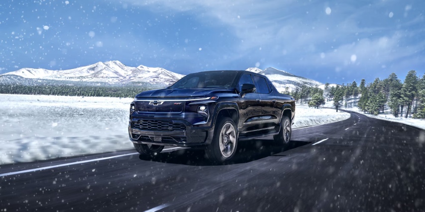 Dark blue Chevrolet pickup driving through a snowy mountain road with snowfall and pine trees lining the route.